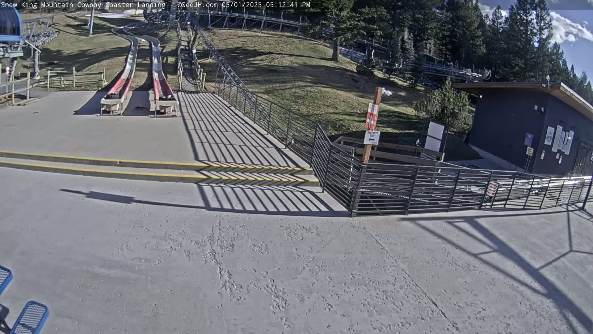 A sunny outdoor scene shows the landing area of a snow coaster with two empty red and white sleds, a small building, and a metal fence.