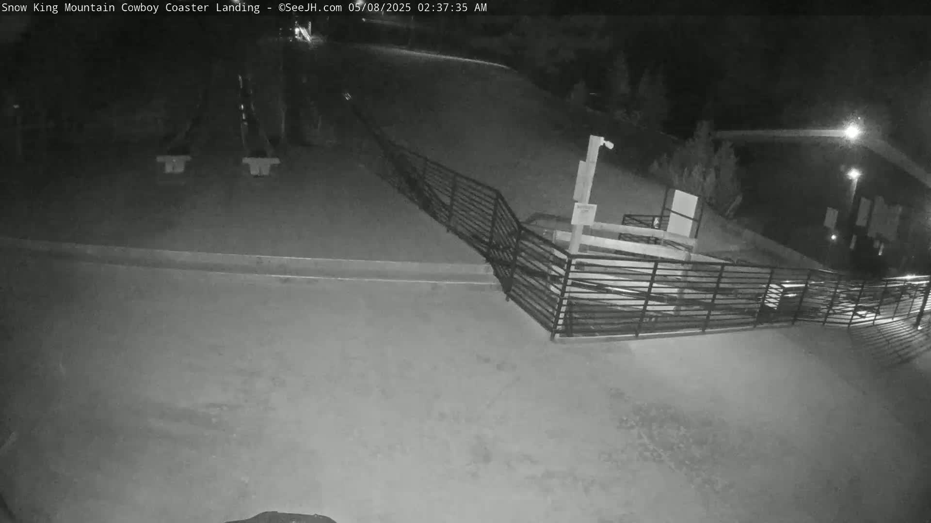 A nighttime, low-light view shows an empty paved area with a metal railing and a dark building in the background.