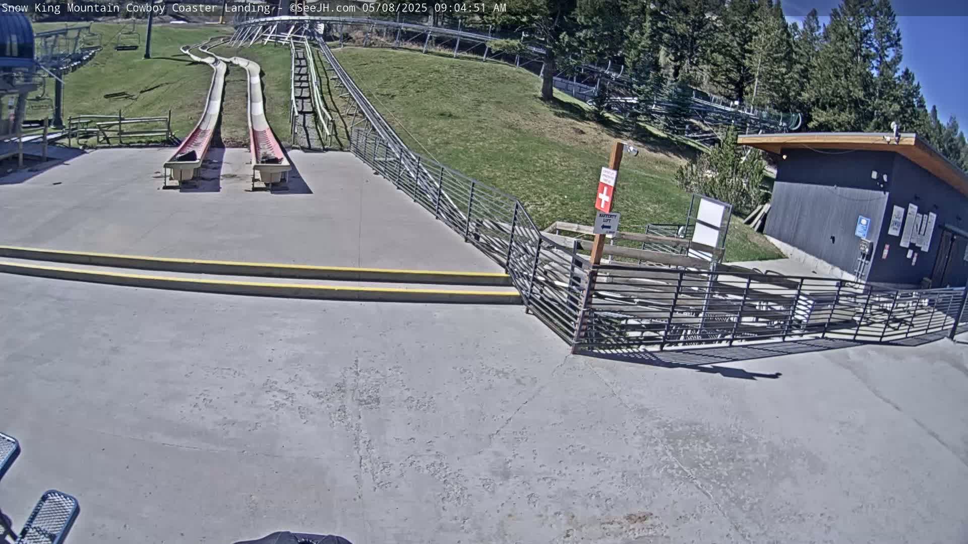 Two empty, side-by-side slides are at the bottom of a hill on a sunny day, near a small building and metal fencing.