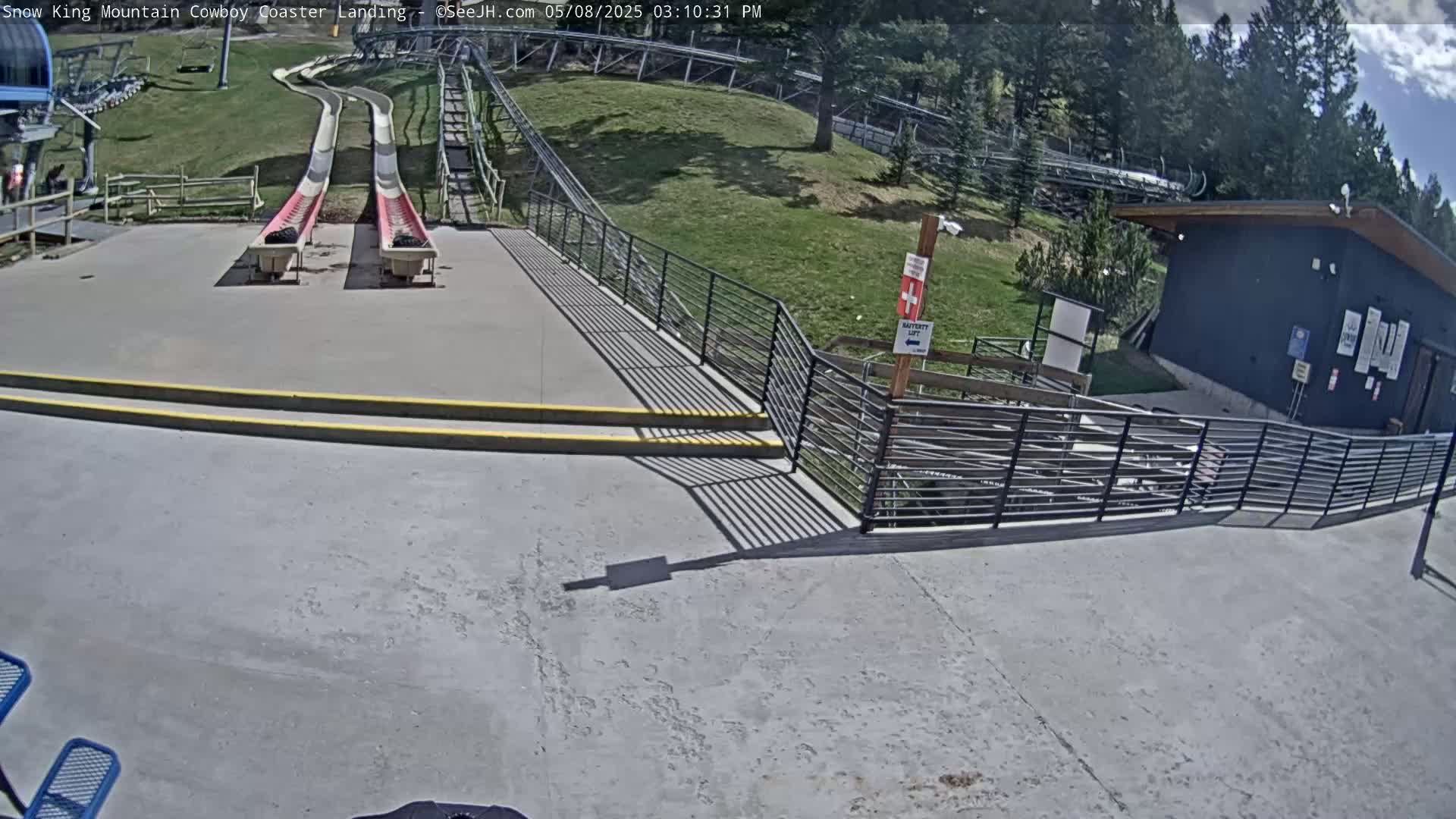 Two red and white coaster slides end at a paved landing area next to a small building under a partly cloudy sky.