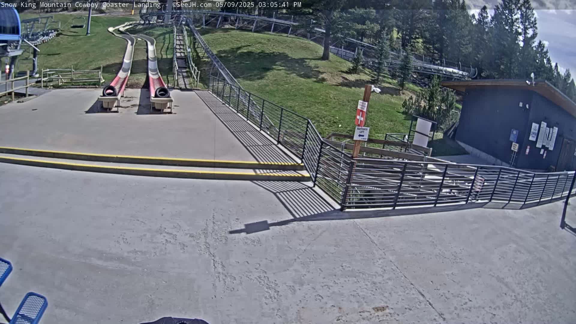 Two long, curved slides end at a paved area with railings, stairs, and a small building, all under a sunny sky.