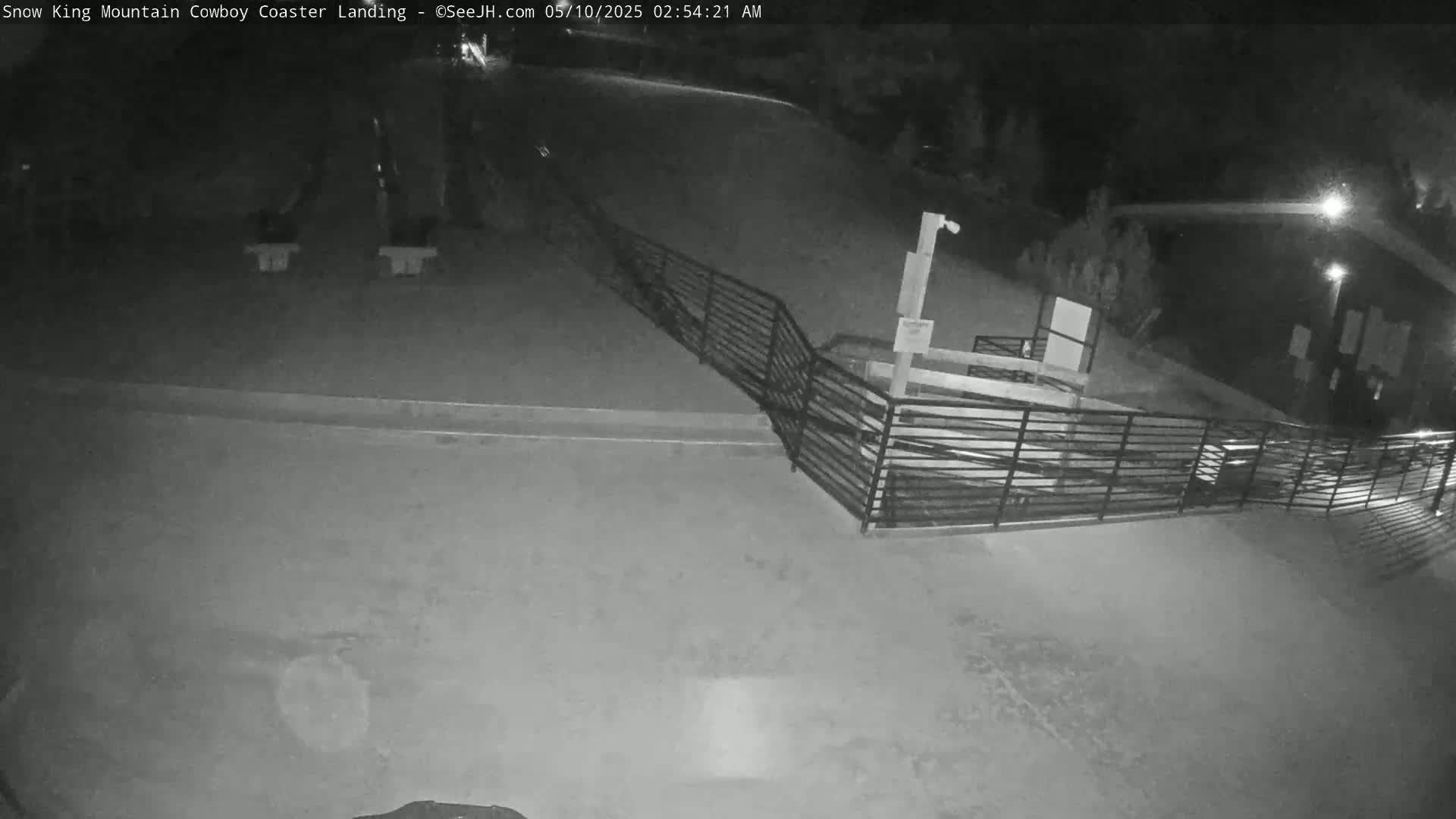 A nighttime, low-light view shows a mostly empty paved area with a metal railing and a building in the background.