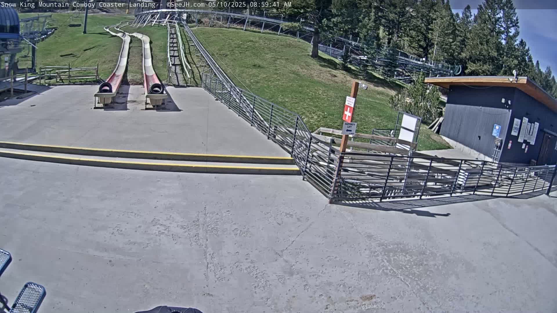 Two empty, red-and-white slides end at a paved landing area next to a small building under a sunny sky.
