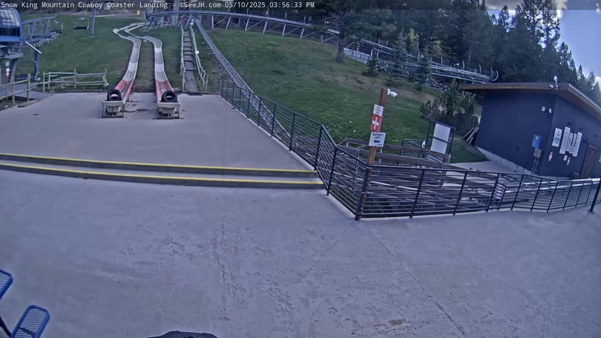 Two empty slide tracks end at a paved landing area next to a small building, all situated on a grassy hillside under a partly cloudy sky.