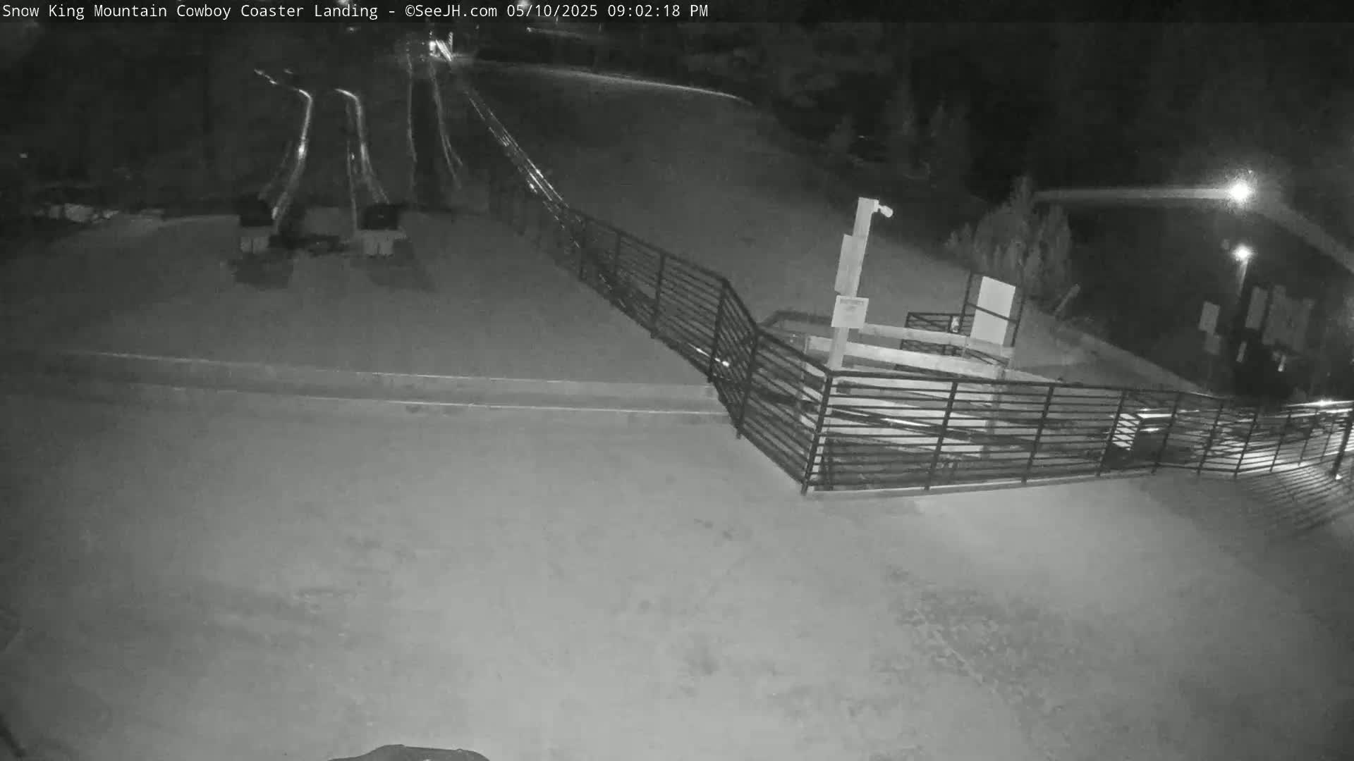 A nighttime, outdoor view shows a paved area with a metal fence, a building, and light streaks suggesting movement.