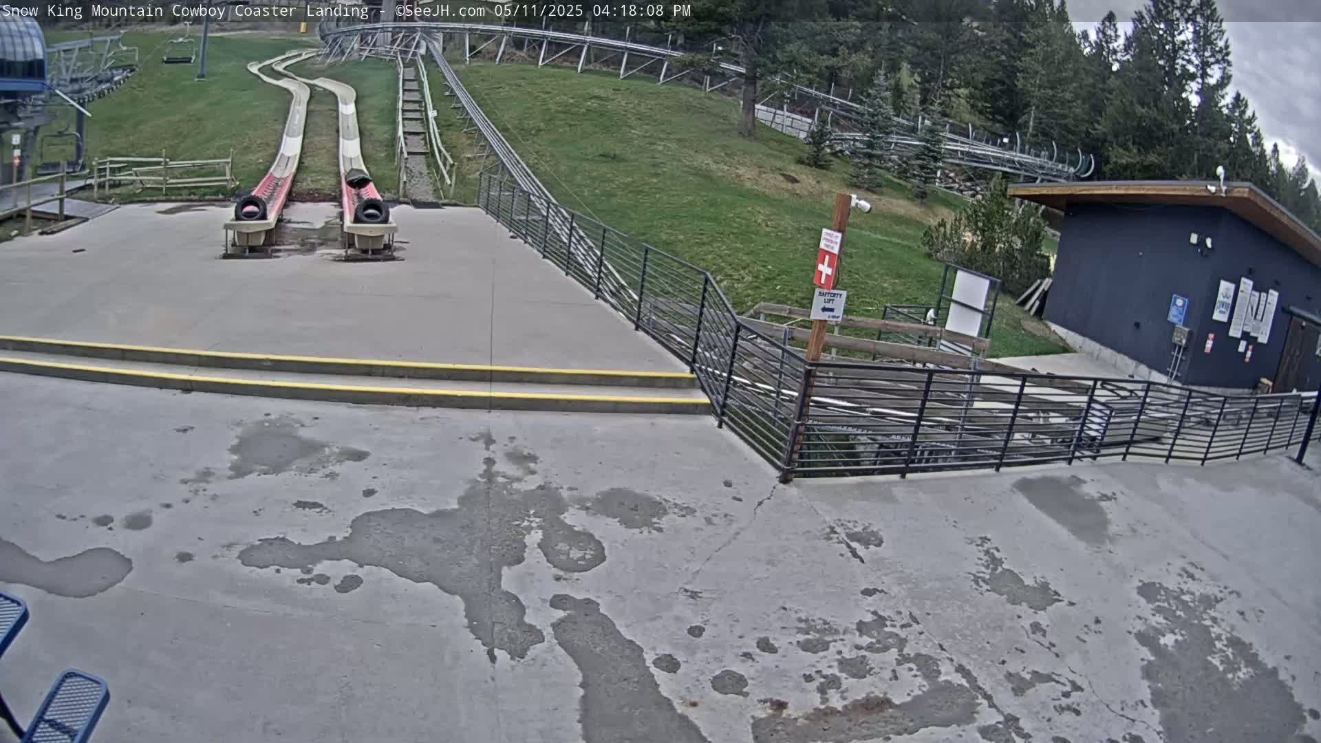 Two long, curving slides end at a concrete landing area next to a small building, all situated on a grassy hillside under an overcast sky.