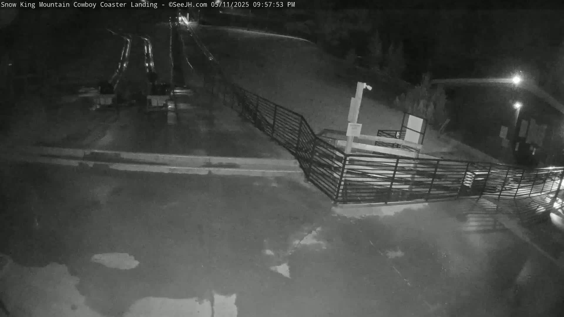 A nighttime, wet, paved area with a metal fence and a building in the background, showing what appears to be a coaster landing area.