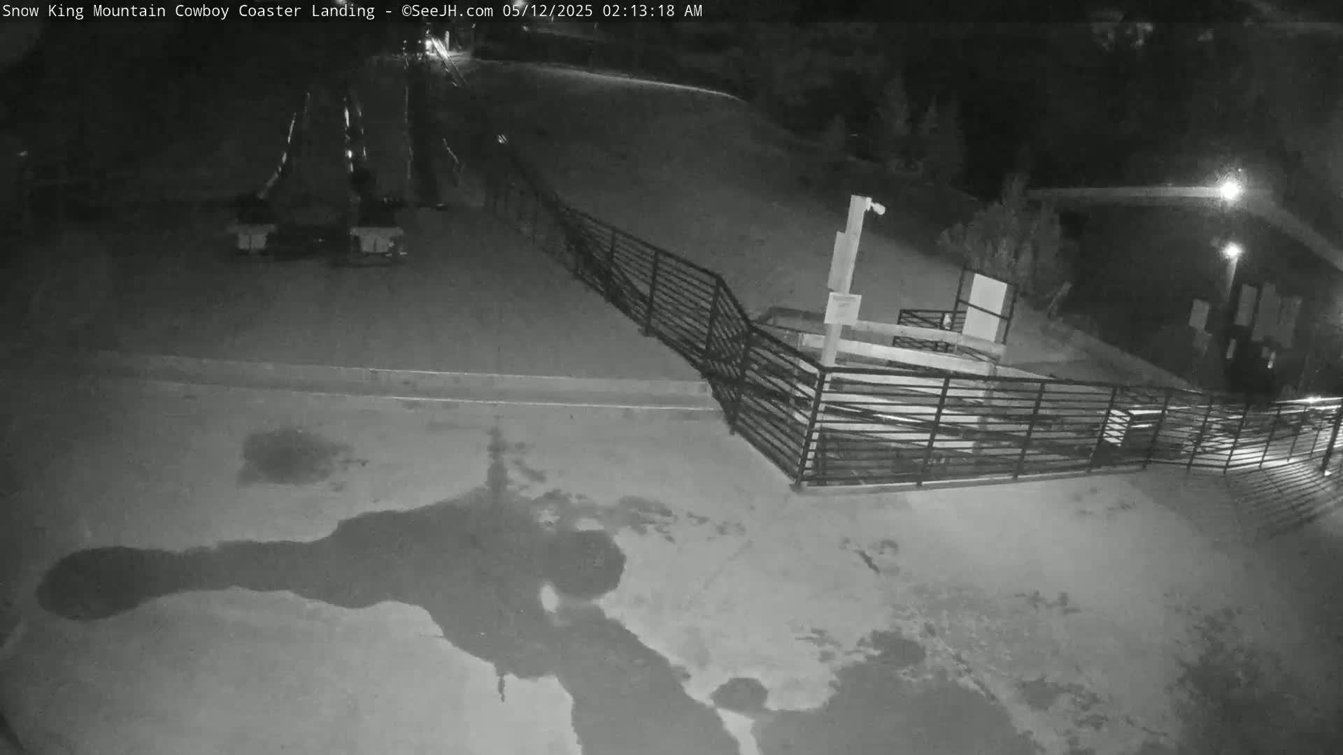 A nighttime, low-light view shows a paved area with a metal fence and some dark puddles, near a building.