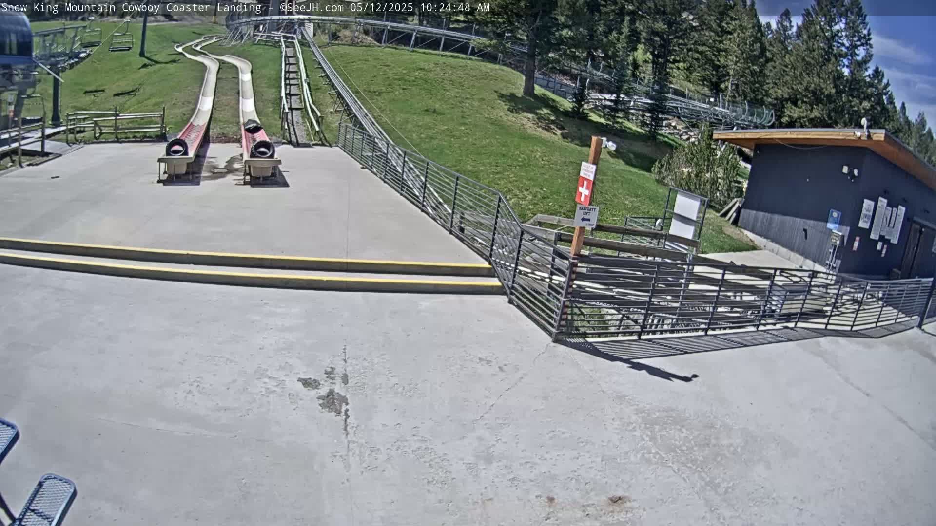 Two empty alpine slides end at a concrete landing area next to a small building, under a mostly sunny sky.
