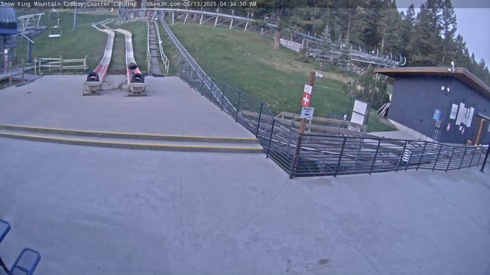 Two empty alpine slides end at a paved landing area next to a small building, in front of a grassy hill with trees under a cloudy sky.