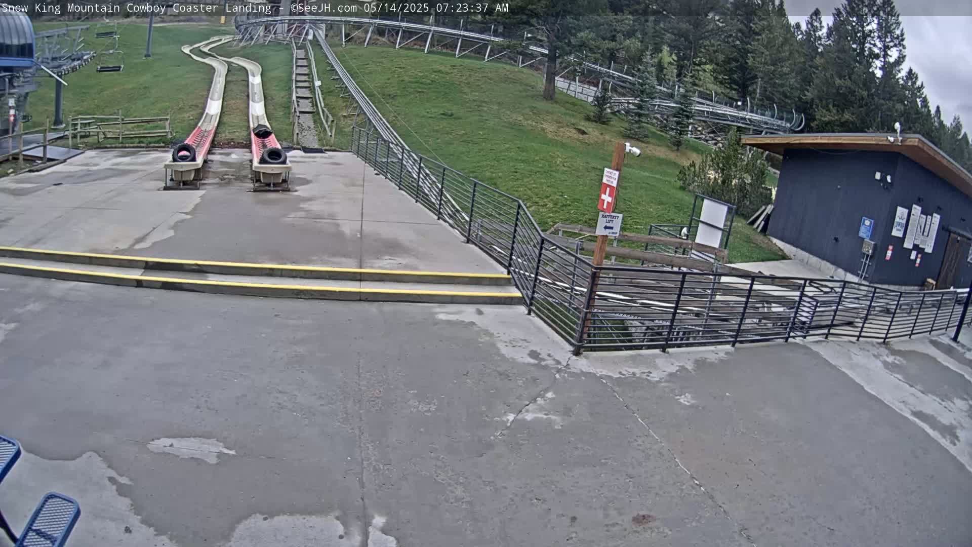 Two empty alpine coasters end at a concrete landing area next to a small building, with grassy hills and trees visible in the background under an overcast sky.