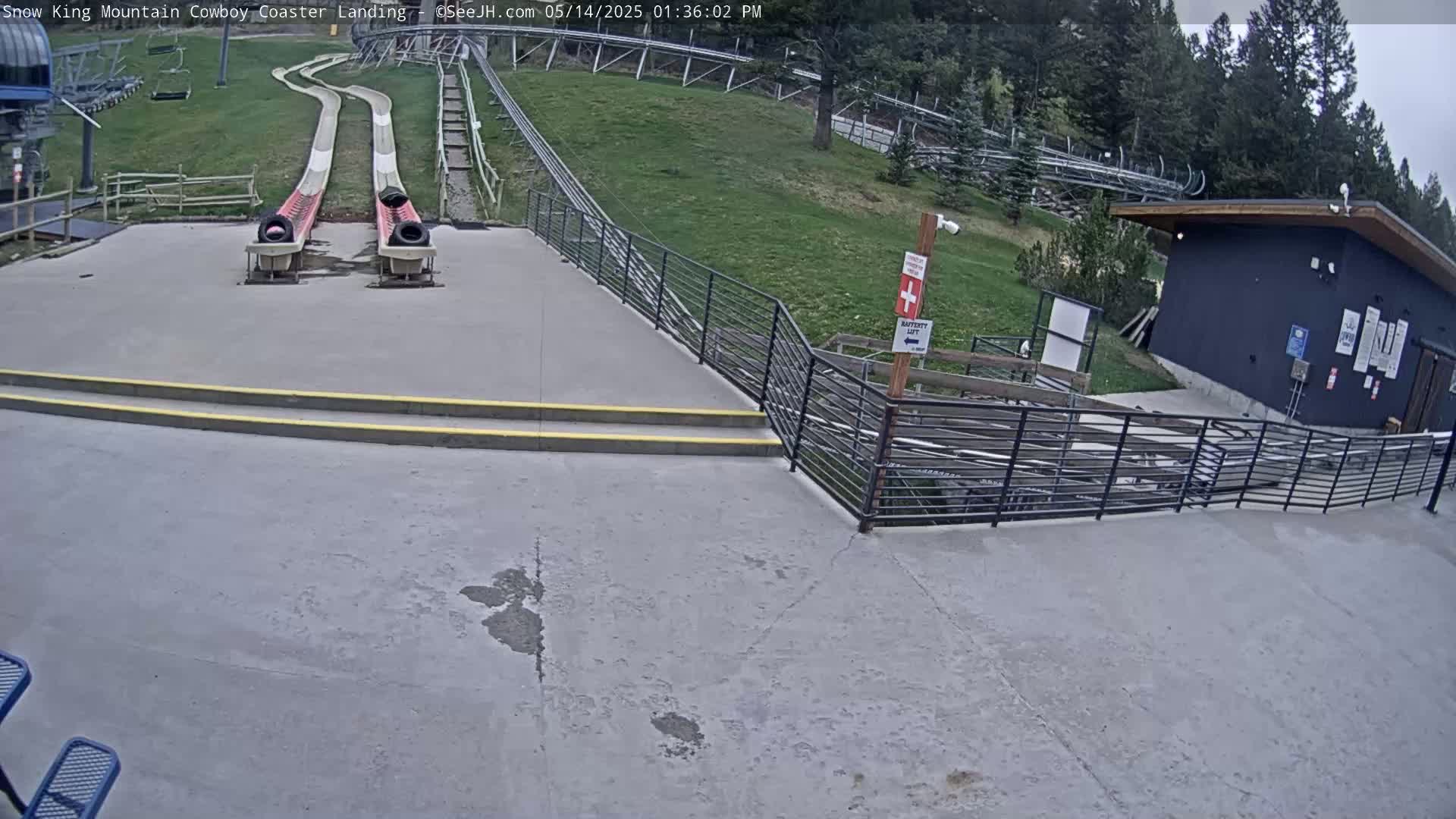 Two empty sleds sit at the bottom of a coaster track, alongside a paved area with stairs and a small building, all set against a backdrop of a grassy hillside and trees under a partly cloudy sky.