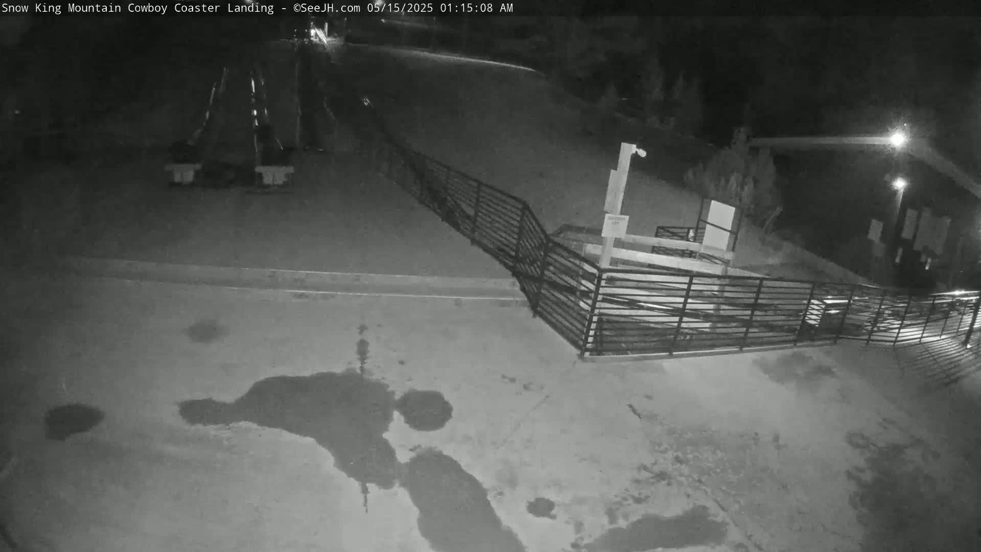 A nighttime, low-angle view shows a paved area with some puddles, a metal railing, and two coaster-like structures in the distance.