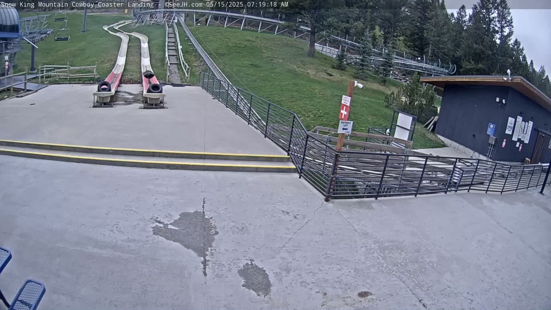 Two empty, side-by-side slides at the bottom of a hill are situated near a small building and railing, on an overcast day.
