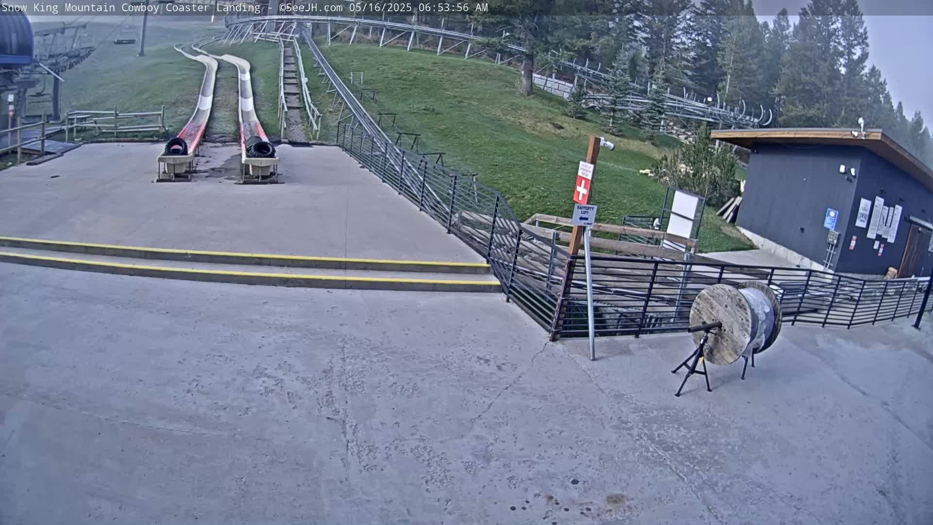 Two empty alpine slides end at a paved landing area next to a small building, under an overcast sky.