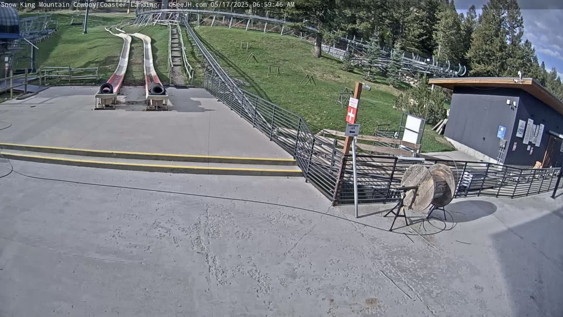 Two long, gently sloping slides end at a paved landing area near a small building, on a sunny day with a grassy hillside in the background.