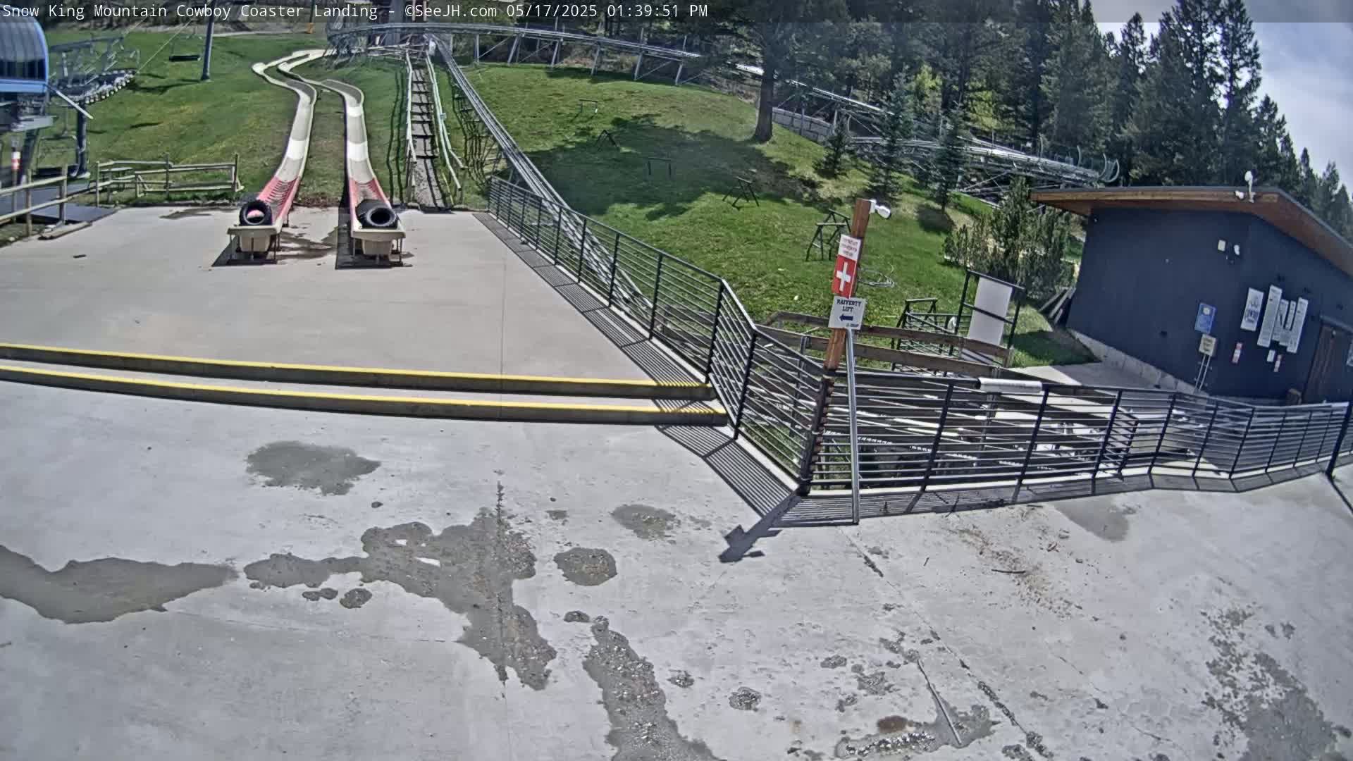 Two empty red and white slides at the bottom of a hill end at a paved landing area with a small building and metal railings in the sunny weather.