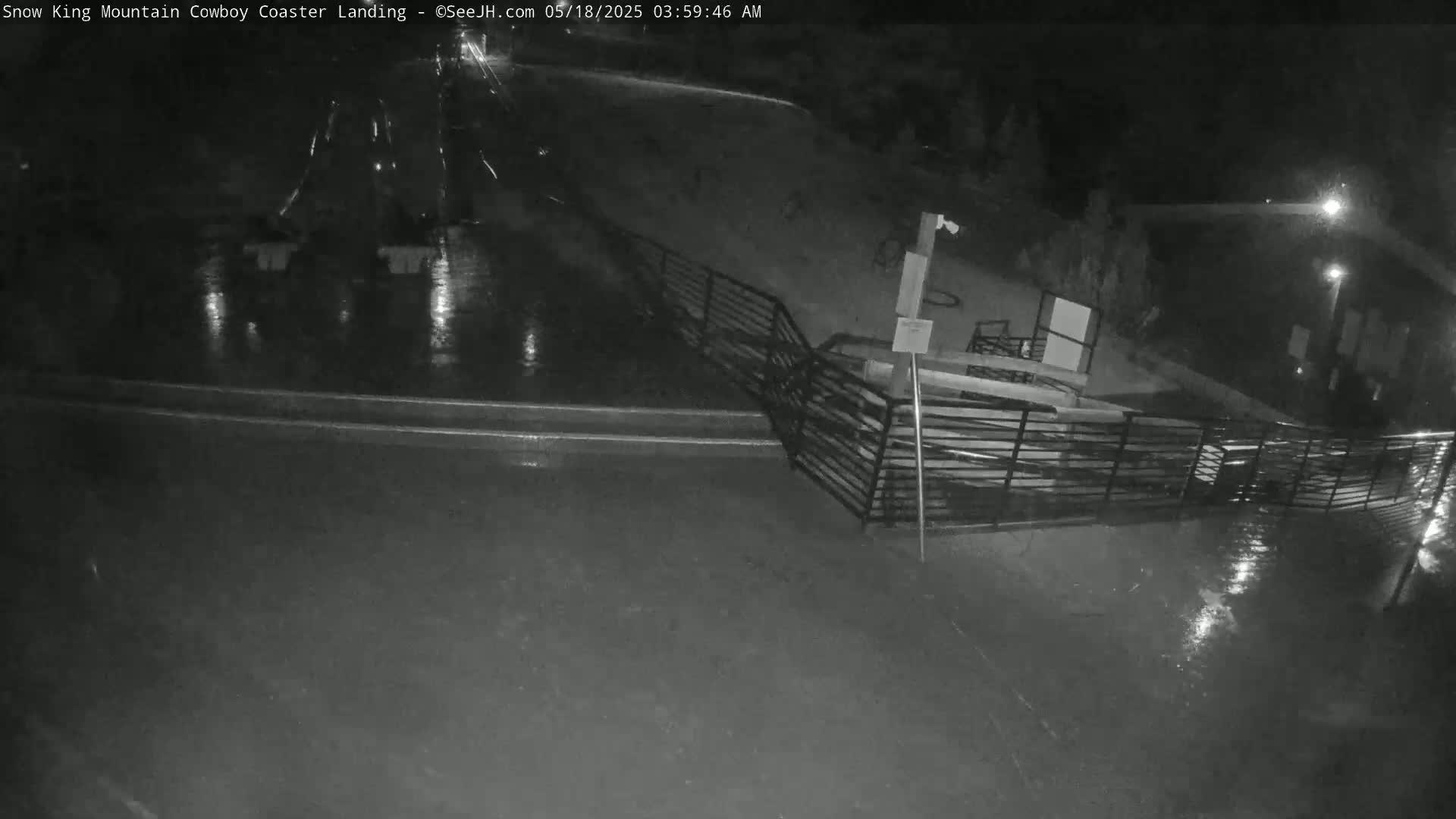 A nighttime, rain-soaked area with a metal railing and a building in the background.