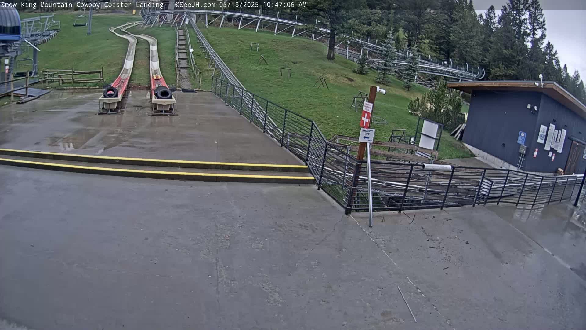 Two empty alpine coasters end at a wet concrete landing area near a small building, with a grassy hillside and trees visible in the background under overcast skies.