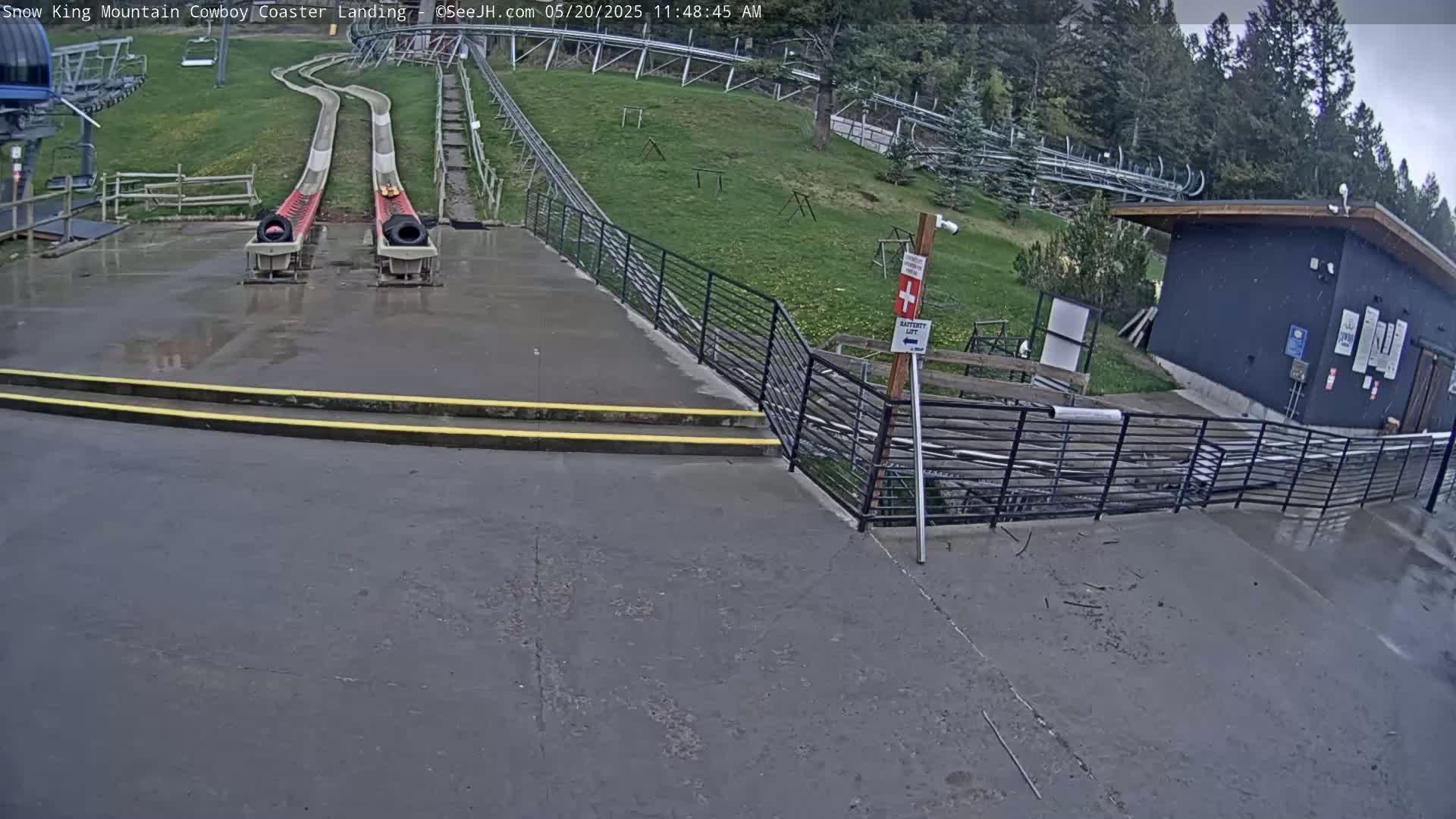 A wet paved area shows two empty alpine coaster tracks leading uphill to a wooded area, with a small building and metal railings nearby.