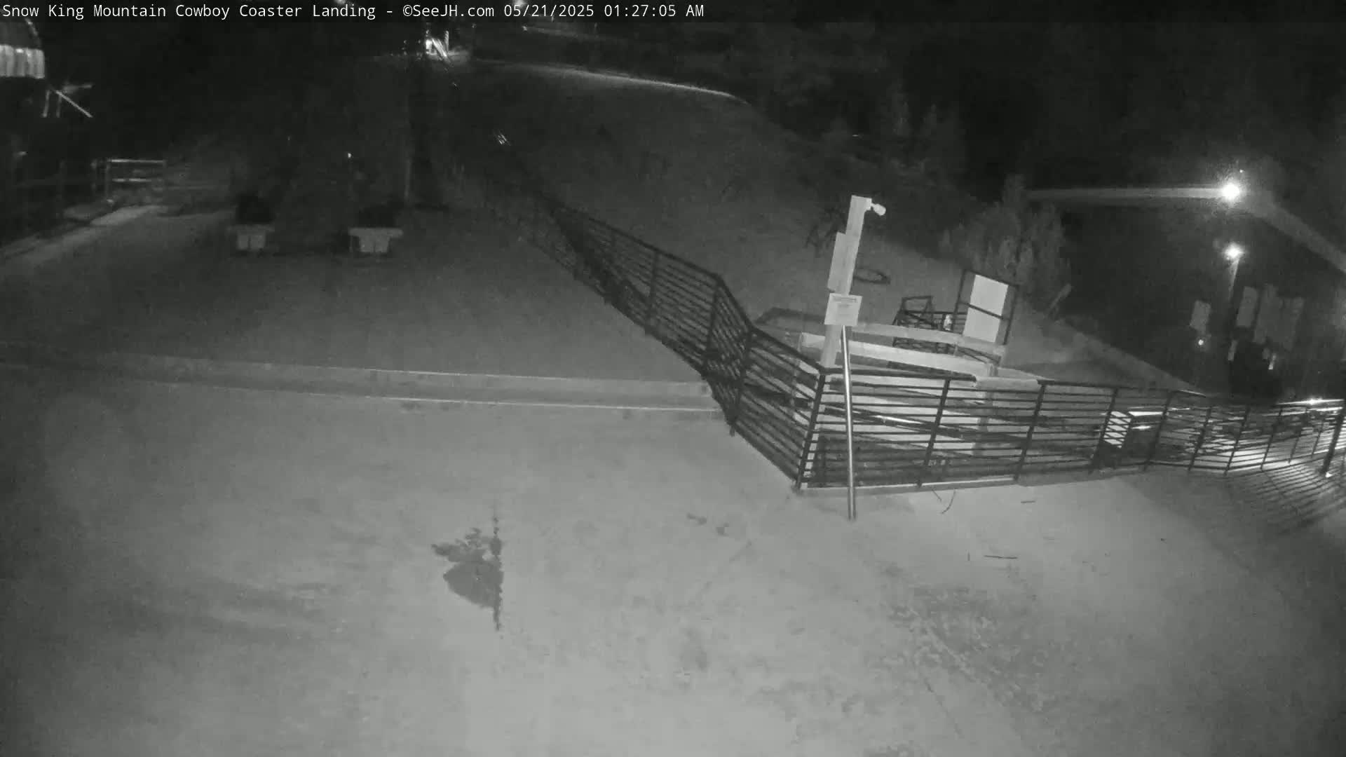 A nighttime, low-light view of a paved area with metal railings, a building, and some indistinct objects in the distance.