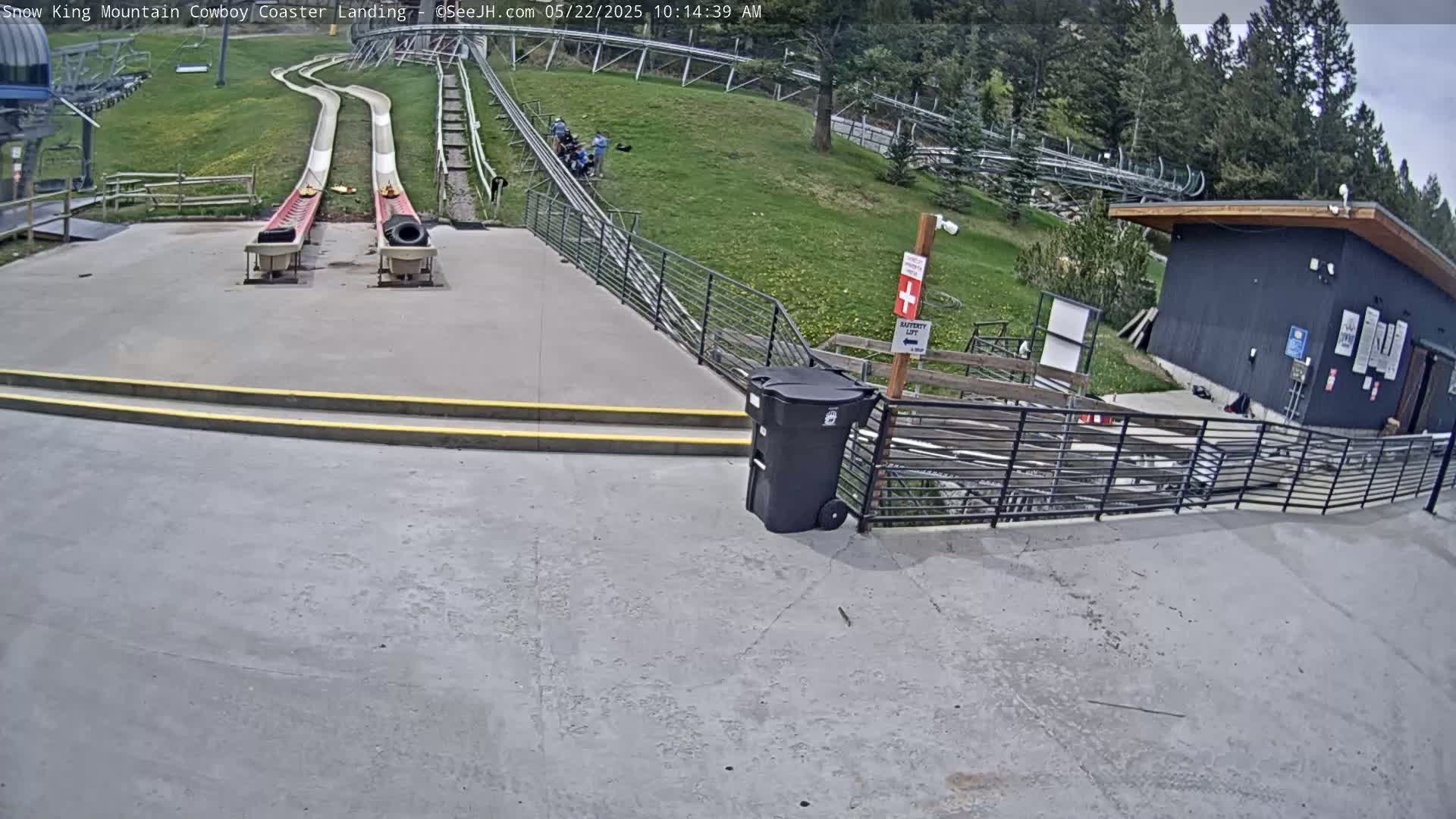 Two empty sleds sit at the bottom of a pair of long, curving slides, near a small building and some people, on a sunny day.