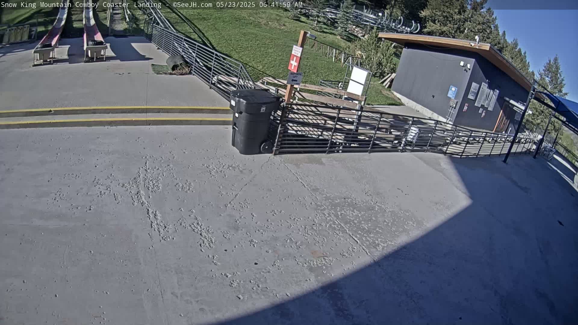 A paved area with a metal fence, a small building, and two empty coaster tracks is visible under sunny conditions.