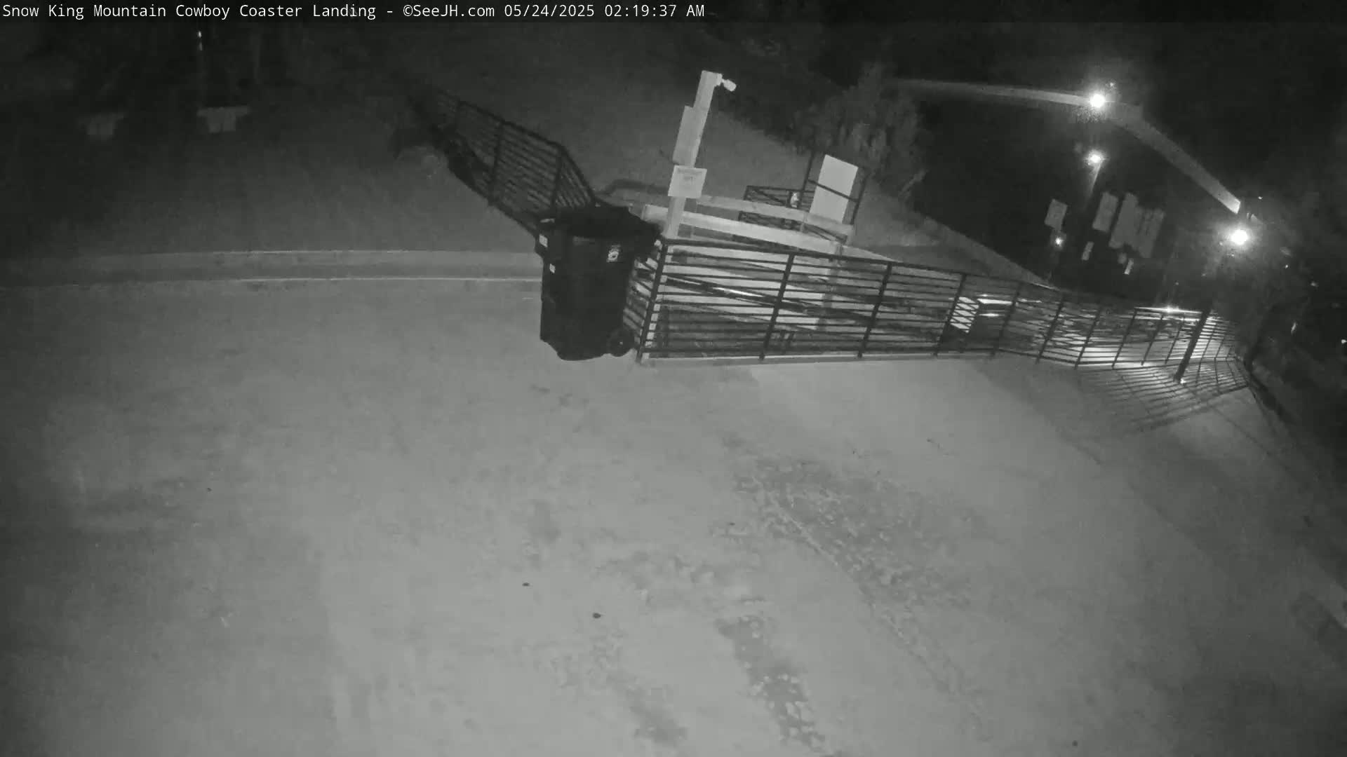 A nighttime, black-and-white view shows a paved area with a metal fence and a trash can near a building, illuminated by artificial light.