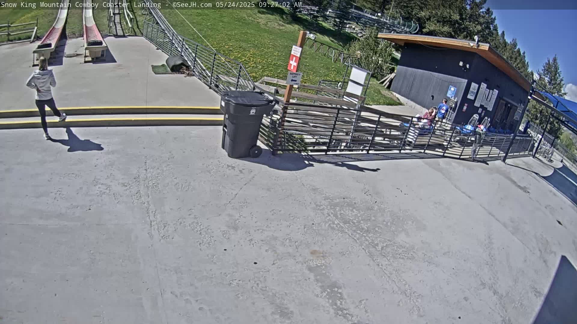 A person walks away from a coaster landing area on a sunny day near a building and a trash can.
