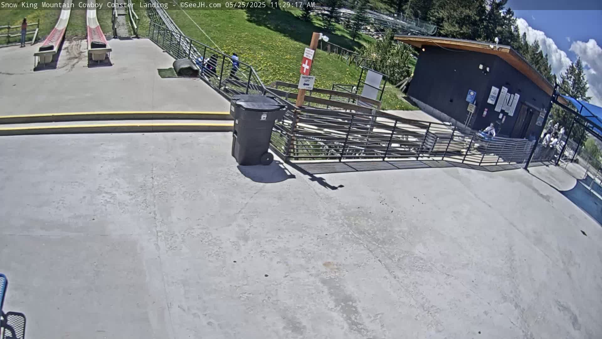 A sunny outdoor scene shows a paved landing area for a coaster with two empty tracks, a small building, a trash can, and a few people.