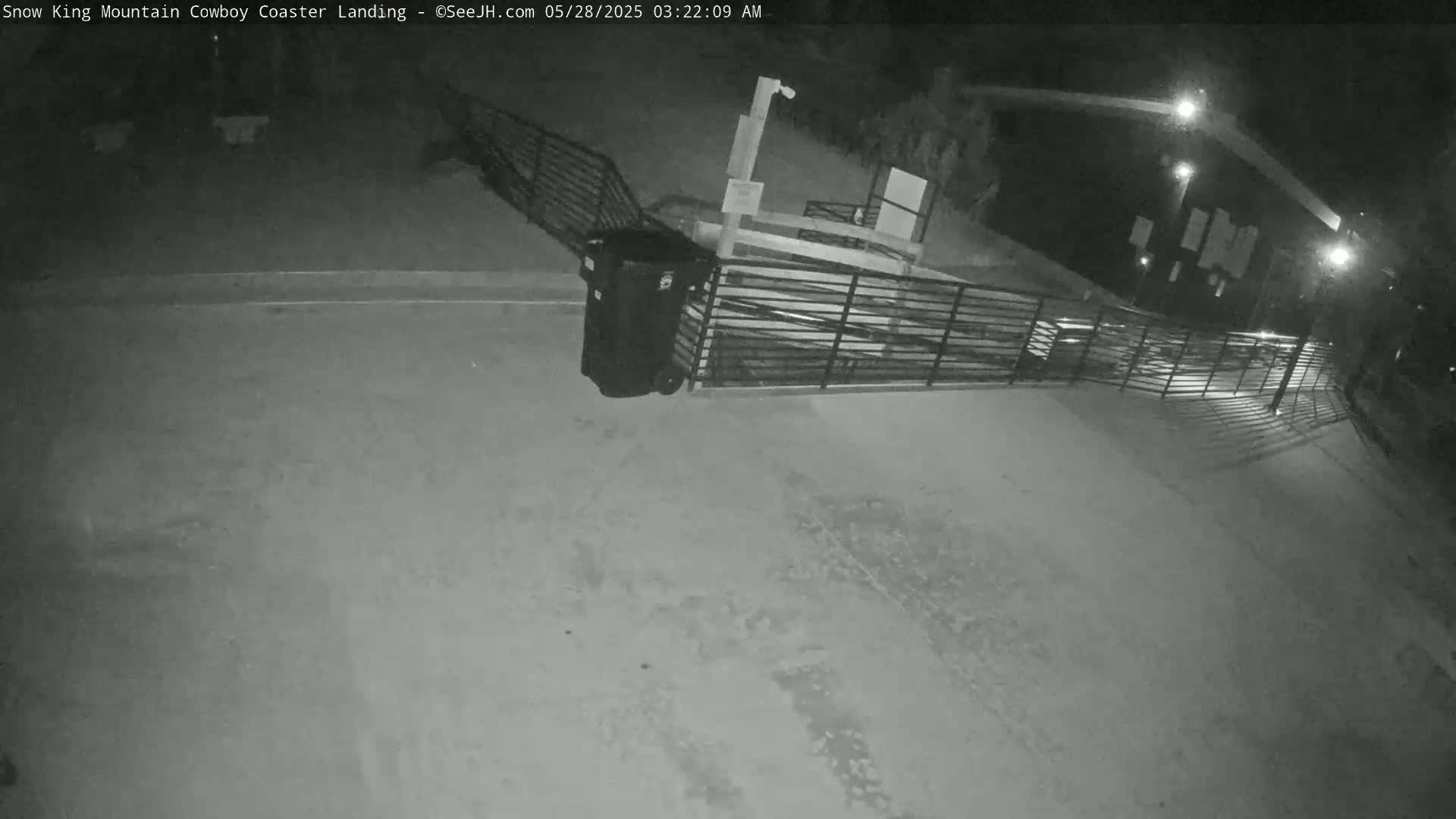 A nighttime, black and white image shows a dark, empty paved area with a metal fence and a large trash can near a building illuminated by artificial light.