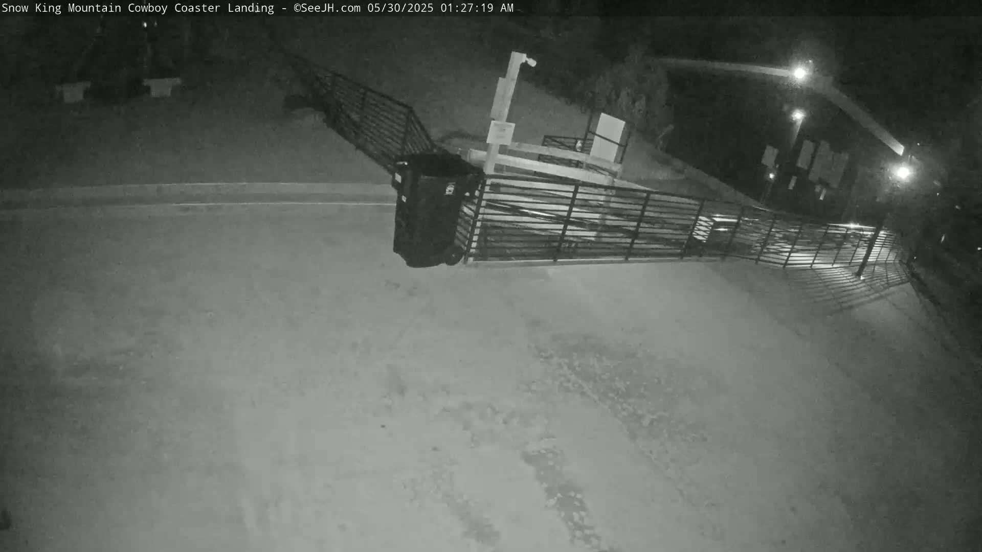 A nighttime, black-and-white security camera view shows an empty paved area with a metal railing and a trash can near a building, under artificial light.