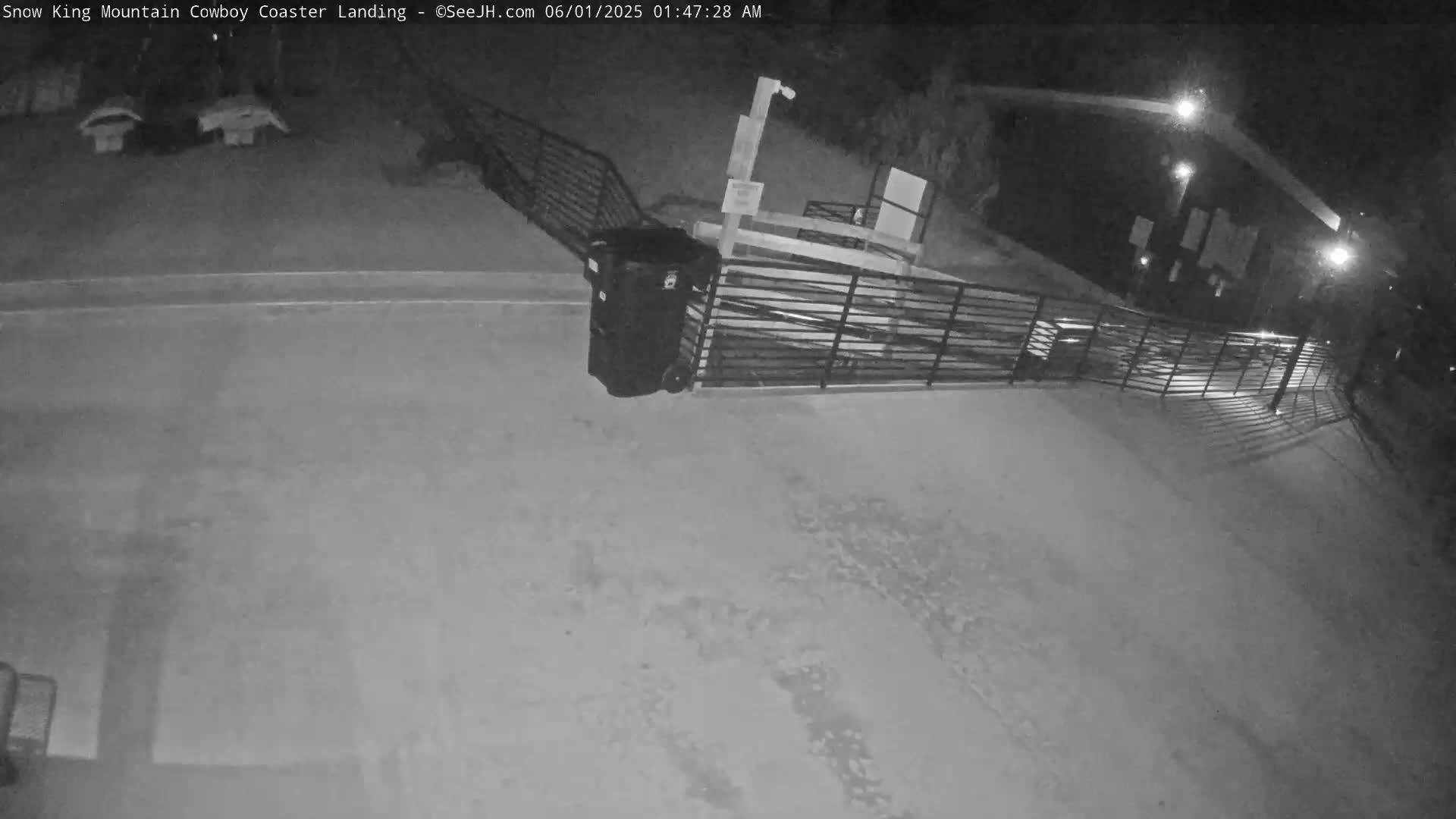 A nighttime, outdoor view shows a paved area with a metal fence and a large trash receptacle near a building.