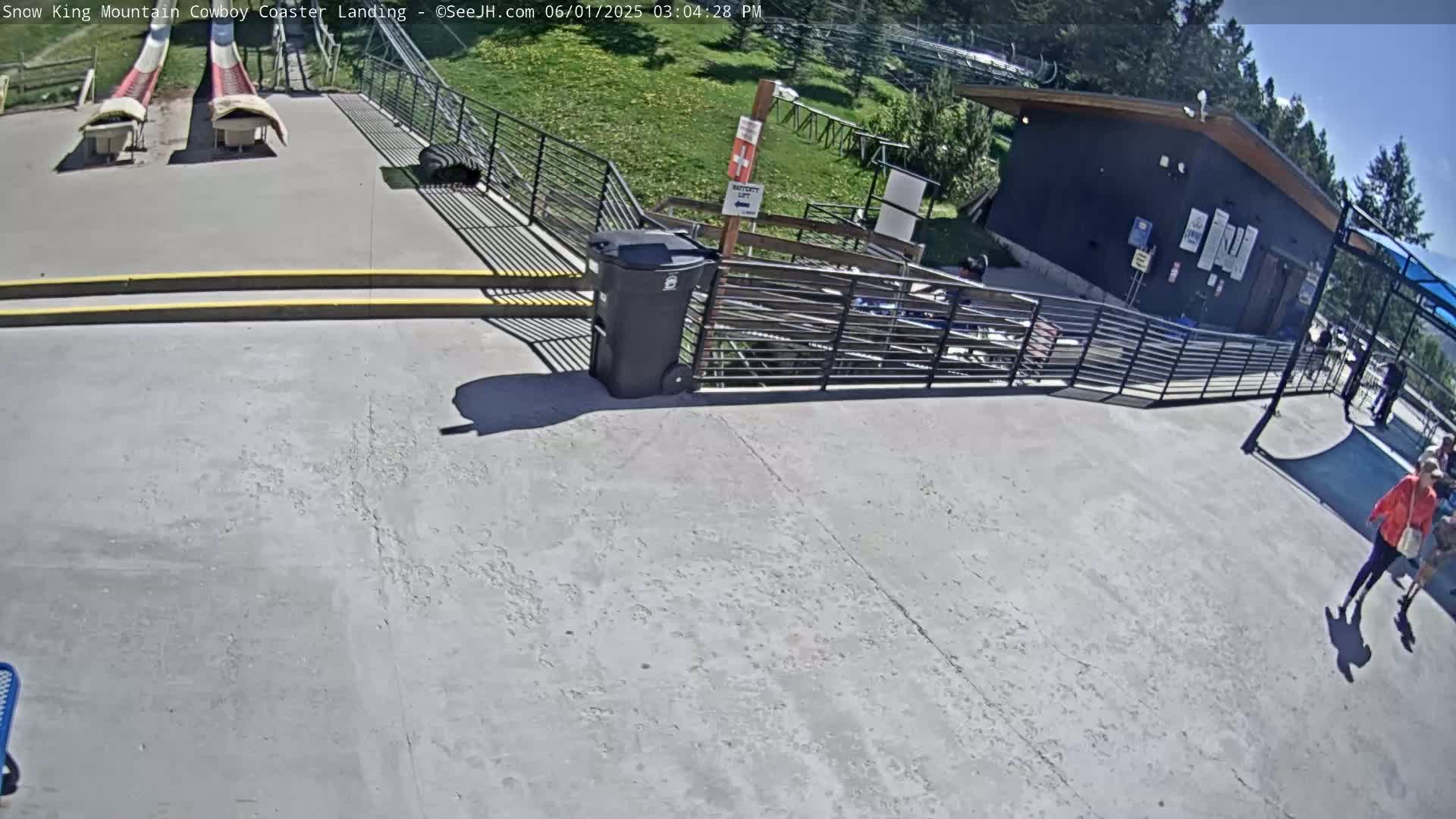 A paved area with two empty, red and white slides, a black building, and a few people walking on a sunny day.