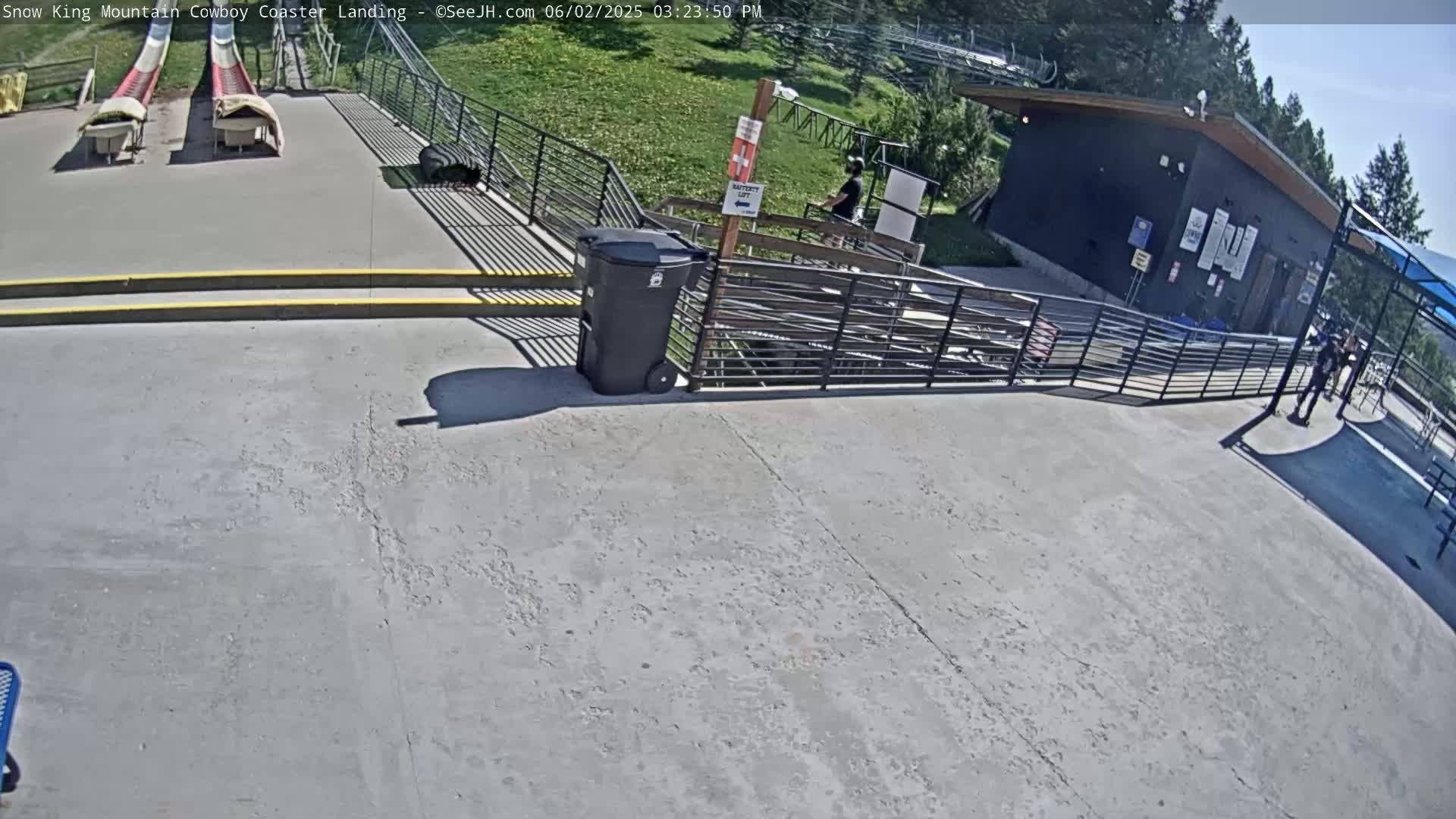 A paved area shows two empty, red toboggan runs, a metal railing, a trash can, and a small building under a sunny sky.