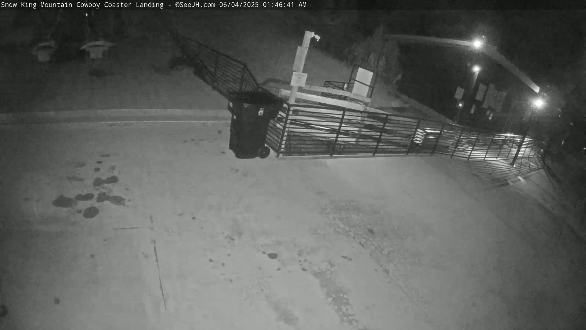 A nighttime, black-and-white image shows a trash can near a metal fence and ramp outside a building illuminated by artificial light.