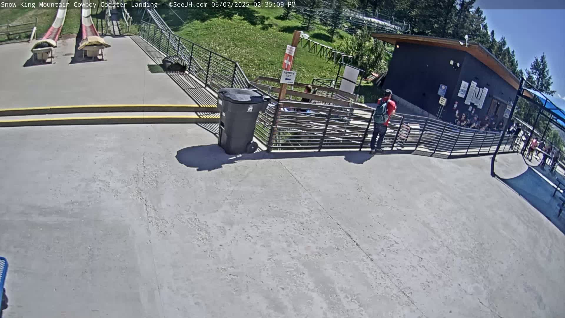 A paved area with two red and white slides, a black trash can, a small building, and several people, under sunny conditions.