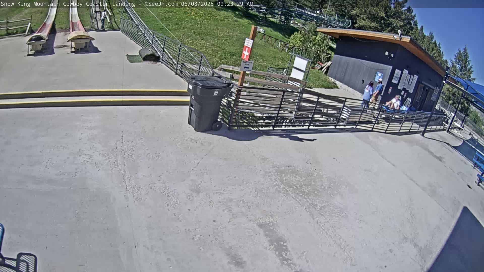 A paved area with two empty roller coasters, a building, some people, and a trash can is shown on a sunny day.