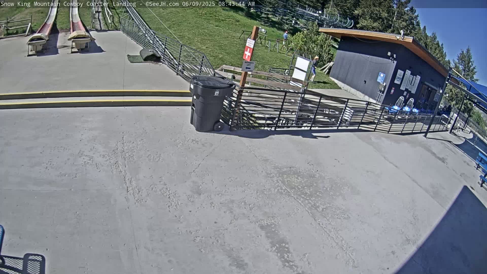 A paved area with two empty coaster tracks, a small building, and a trash can is visible on a sunny day.