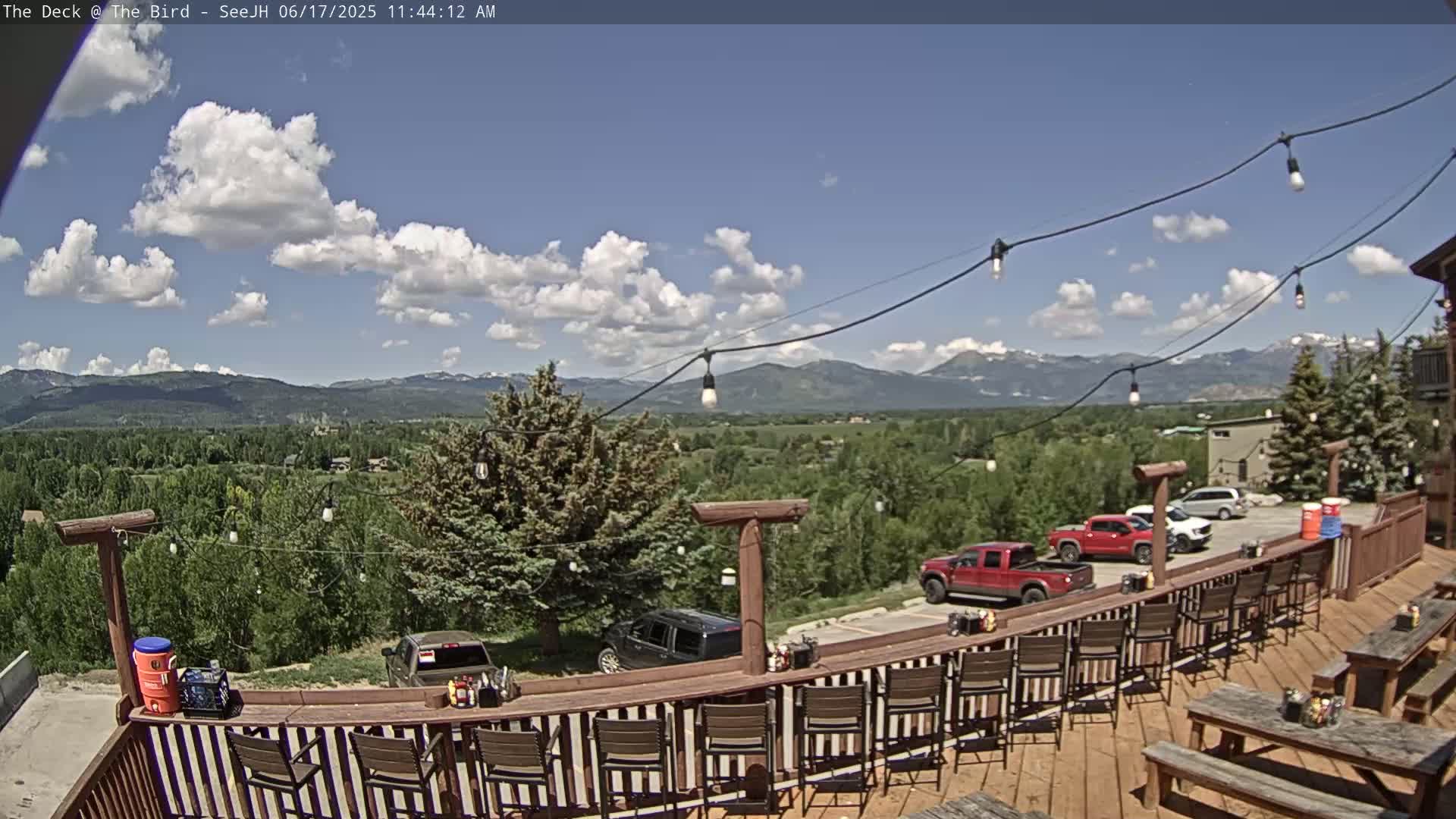 An outdoor deck with a bar and picnic tables overlooks a valley and mountains under a partly cloudy sky.