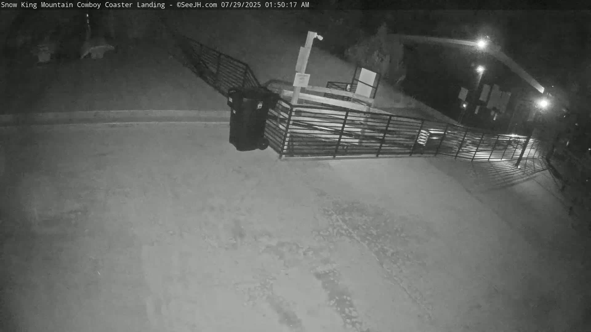 A nighttime, snow-covered area shows a metal fence near a dumpster, with a building visible in the background under artificial light.