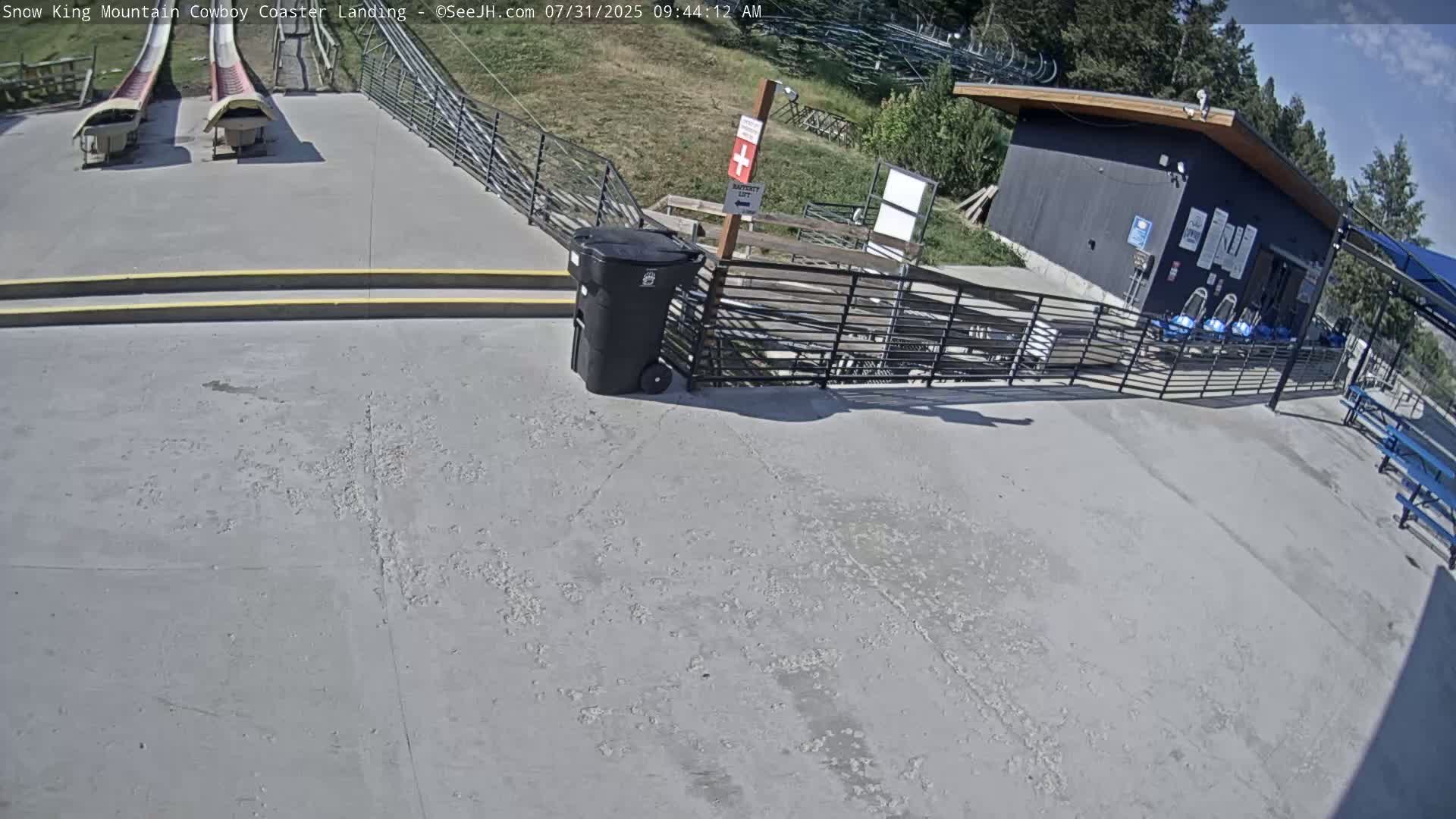 A paved area shows two empty coaster tracks, a small building, a trash can, and a metal fence under a mostly sunny sky.