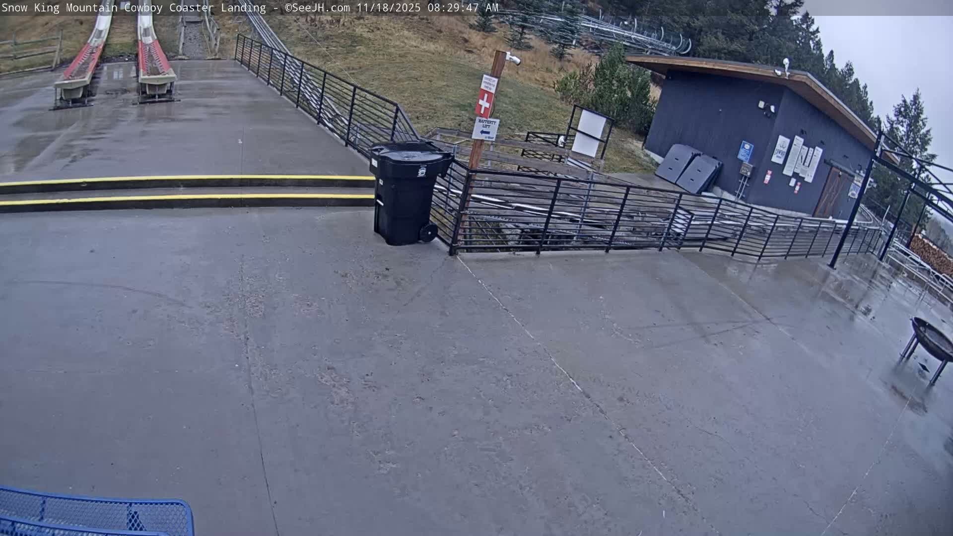 A mostly sunny outdoor scene shows a paved area with two coaster tracks, a small building, and a few people sitting on a bench.