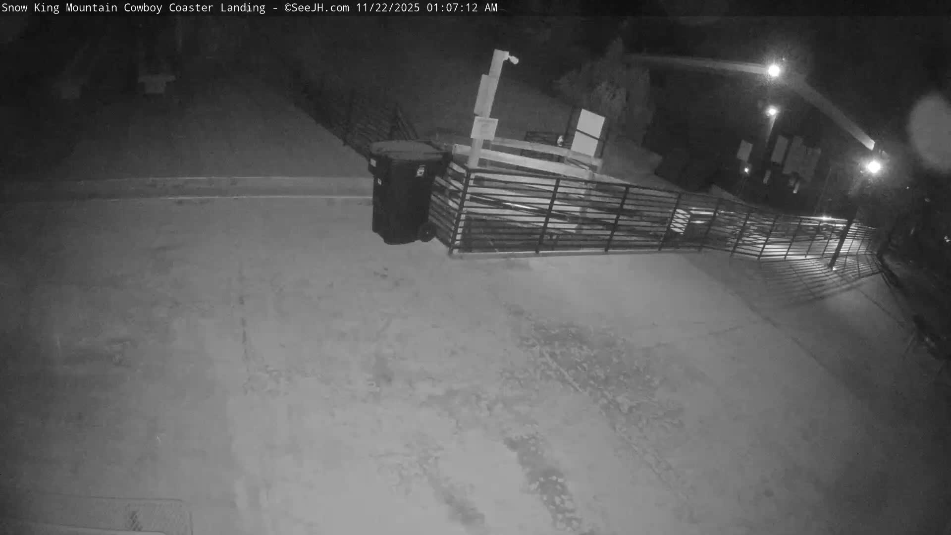 A monochrome image taken at night depicts an outdoor area with a ground partially covered in thin snow and tire tracks, featuring a trash can, fencing, and brightly lit structures under clear, cold conditions.