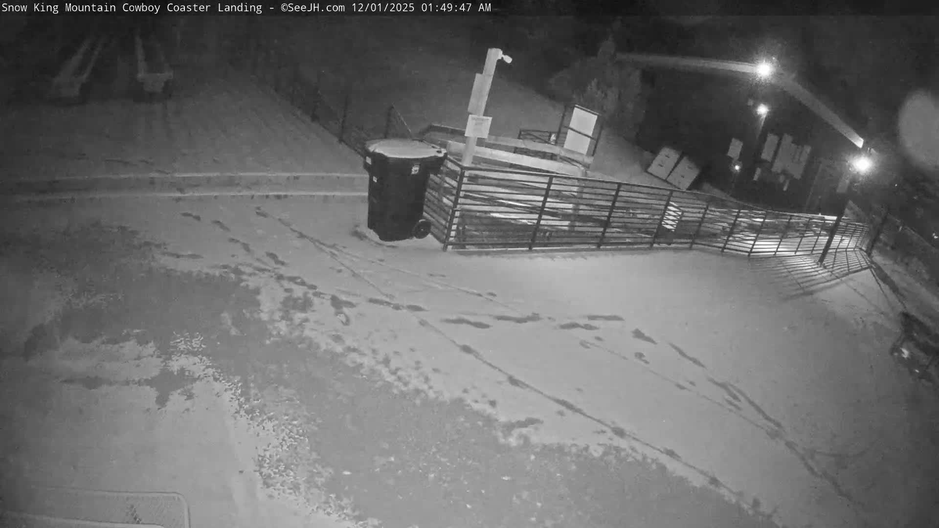 The scene depicts a wintry night outdoors, with patchy snow covering the ground marked by footprints, illuminated by bright artificial lights, and featuring a large trash bin next to a metal fence and background structures.