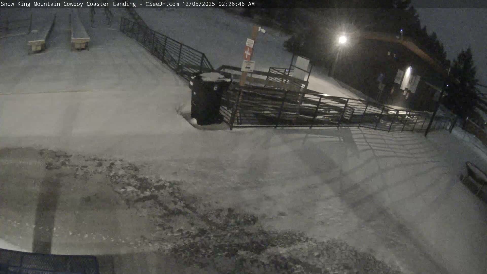 A snow-covered outdoor landing area featuring parallel tracks, fences, a trash bin, and a distant building is illuminated by artificial lights under clear, cold night conditions.