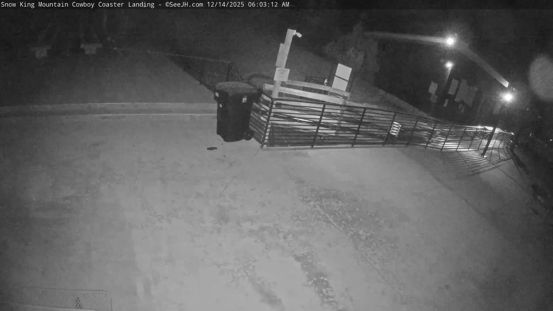 A snow-covered outdoor landing area featuring parallel tracks, fences, a trash bin, and a distant building is illuminated by artificial lights under clear, cold night conditions.