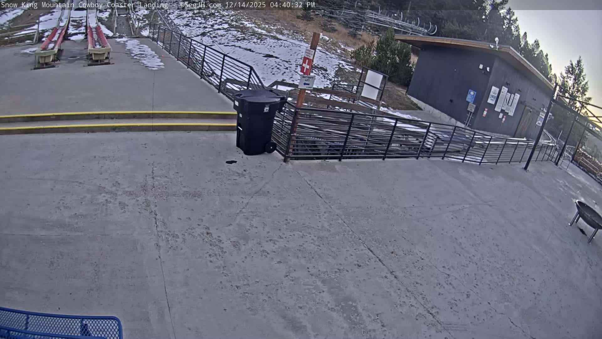 A snow-covered outdoor landing area featuring parallel tracks, fences, a trash bin, and a distant building is illuminated by artificial lights under clear, cold night conditions.