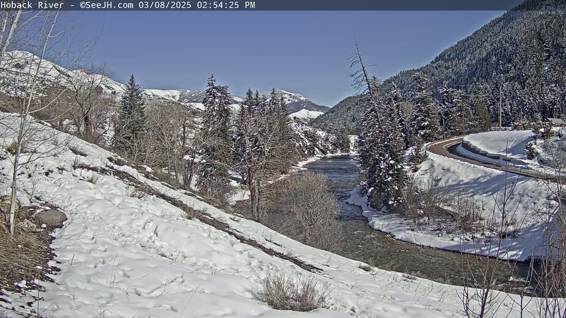 A snow-covered landscape reveals a river winding through a valley under a clear blue sky, with evergreen trees lining the riverbanks and a road curving alongside.