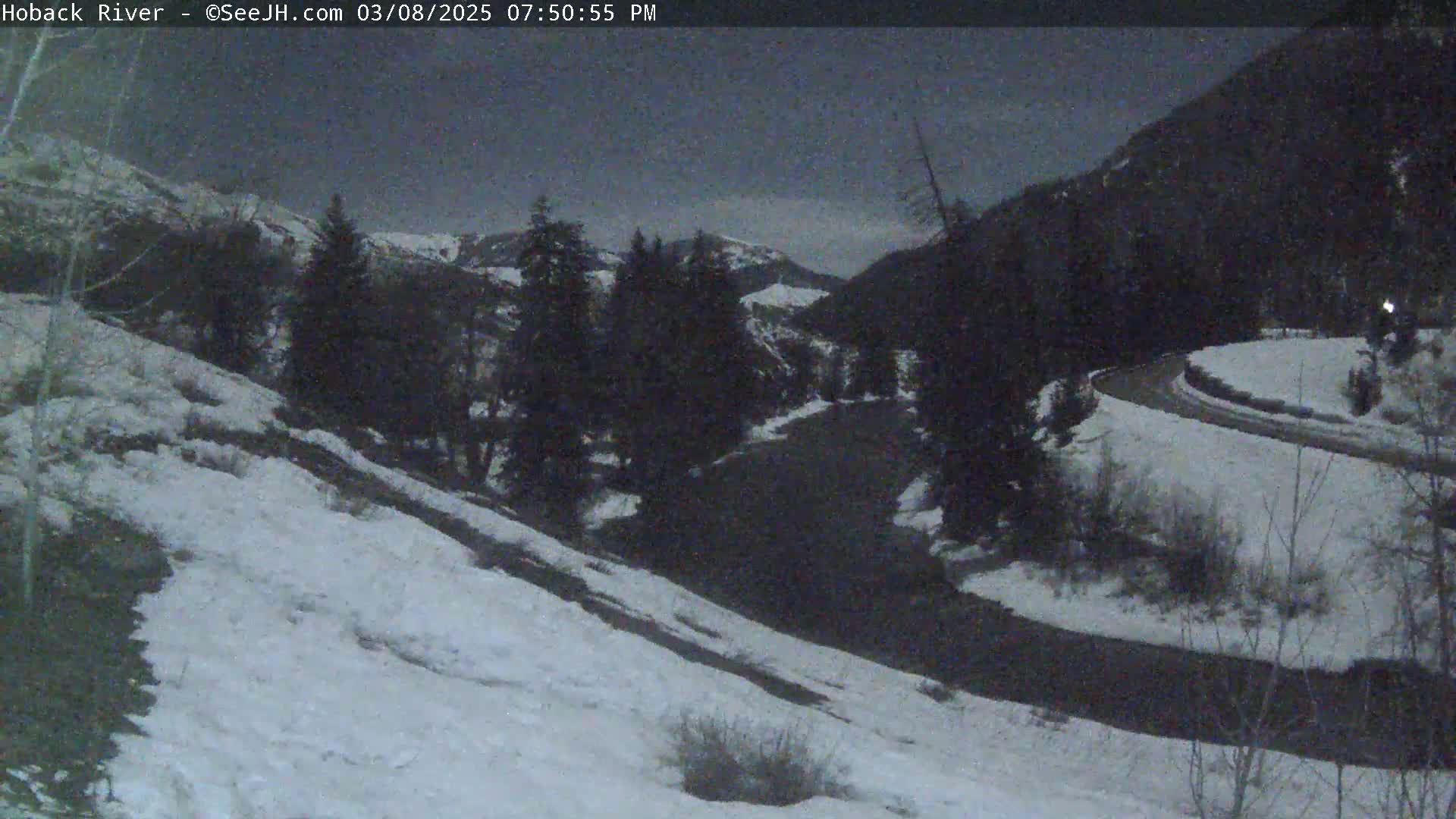 A snow-covered hillside overlooks a dark, winding road that curves through a valley of dark evergreens under a twilight sky.
